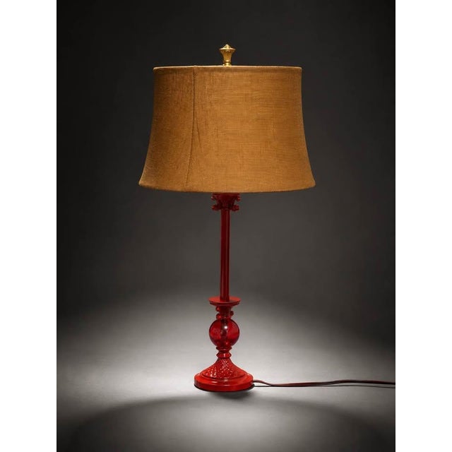 A vintage-style red glass table lamp with a glossy decorative base and a warm burlap-style fabric shade. The bold red base...