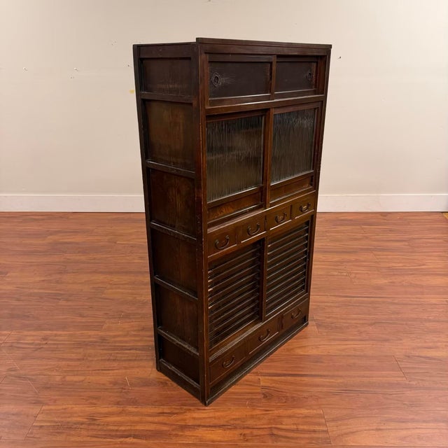 A richly patinated Japanese wooden storage cabinet featuring textured glass sliding doors, staggered display shelves, and...