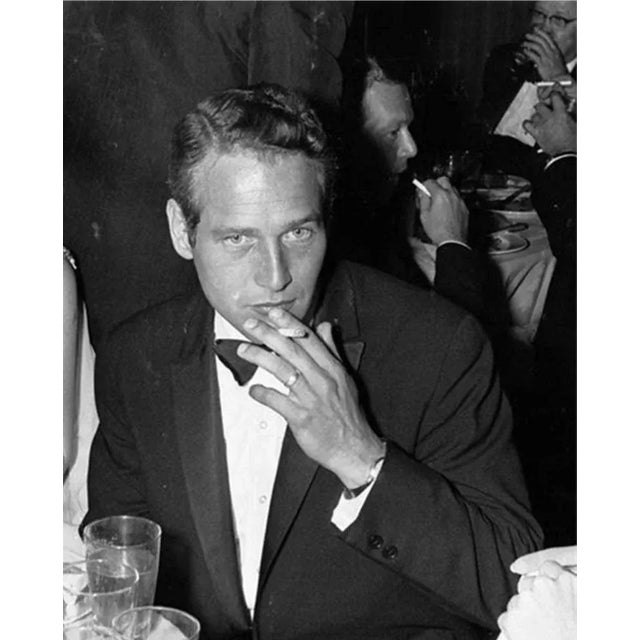 American actor Paul Newman is photographed at a dinner with a cigarette just brought to his lips. It is a carefree, candid...