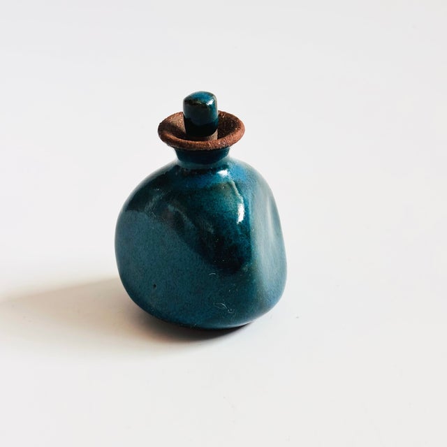 A small vintage studio pottery perfume bottle. Triangular shape with a dark teal glaze. A matching dauber is glazed at...
