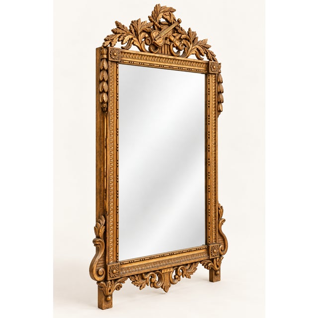 An ornate gilded mirror crowned by a sculptural violin-and-foliage crest. Carved ornament continues along the frame and...