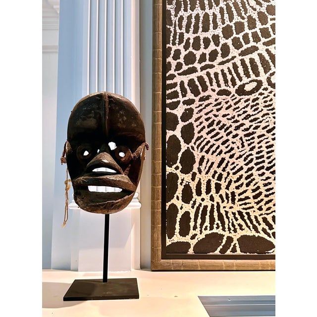 A Guere wood mask with paint and fiber remnant from We people displayed on a metal stand. The We people (also known as the...
