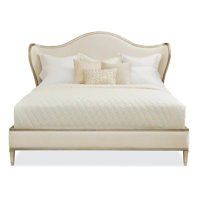 An Upholstered King Bed with a serpentine headboard and curved wings. The exposed carved wood rail is finished in radiant...