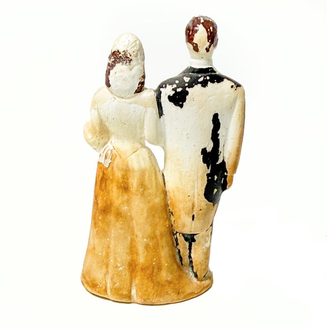 A wonderfully timeworn vintage wedding cake topper featuring a bride and groom standing arm in arm, with beautifully aged...