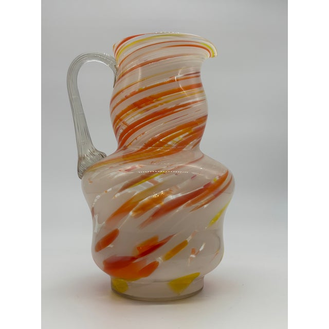 Vintage Italian blown glass pitcher features swirled colors of orange, red, yellow, and white, and has a clear twisted...