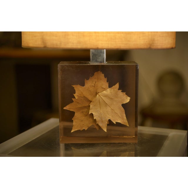 A 1970s table lamp composed of a cylindrical fabric shade and a transparent resin base. The "inclusion" base features a...