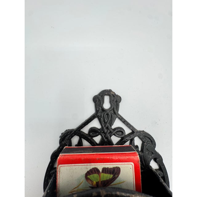 Antique cast iron wall-mounted match holder with ornate scrollwork and a black painted finish showing authentic wear from...