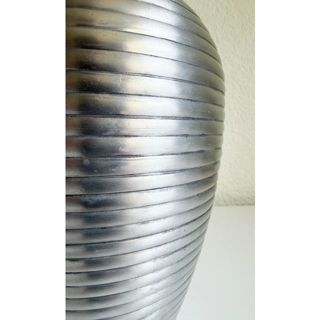 An Art Deco-style ribbed vase crafted from pewter-toned silver metal. It features a sleek, tapered ovoid profile with a...