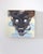 The artwork depicts a striking mask with exaggerated features and luminous eyes, set against a soft, abstract background....