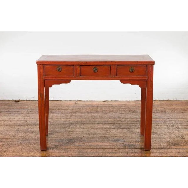 A Chinese Qing Dynasty period table from the 19th century with reddish orange lacquer, three drawers, carved spandrels and...