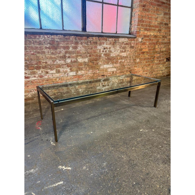 Minimalist brushed bronze coffee table with a thick glass top. Clean rectilinear frame with warm patina on the metal and...