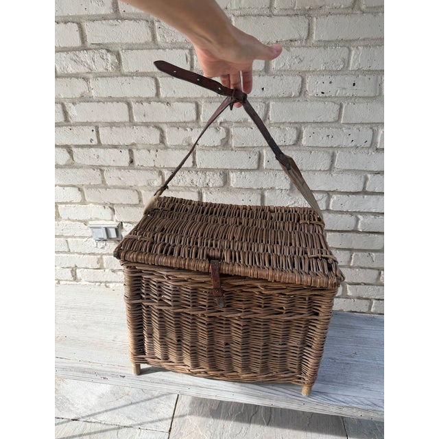 This vintage wicker fishing basket features a rustic hinged lid and a sturdy leather strap for carrying.