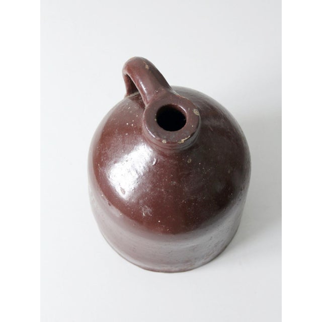 An antique stoneware jug with a rich brown glaze, rounded body, narrow neck, and gracefully curved handle. The deep,...