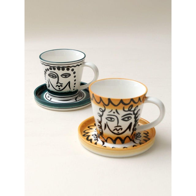 Hand-painted ceramic coffee cup and saucer, featuring abstract human face motifs rendered in line art. The design is...