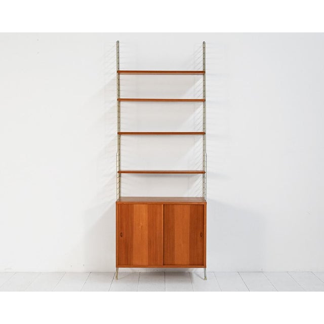 "String" bookcase in teak and lacquered metal with a suspended cabinet with sliding doors and adjustable shelves. String...