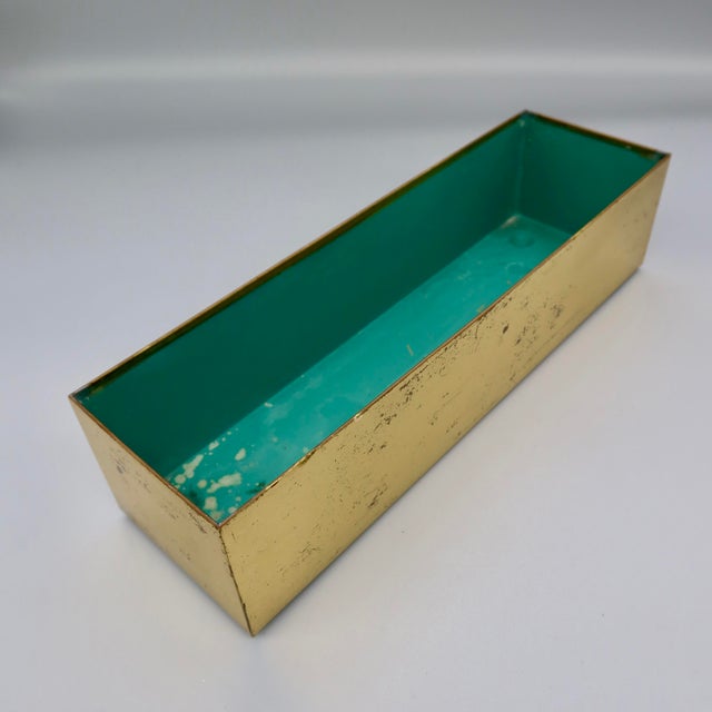 A vintage mid-century modern rectangular box in a brass finish, with a teal-toned interior lining and visible riveted seam...