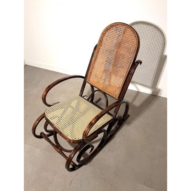 Elegant antique bentwood rocking chair featuring a sculptural dark-stained wooden frame and traditional woven cane seat...
