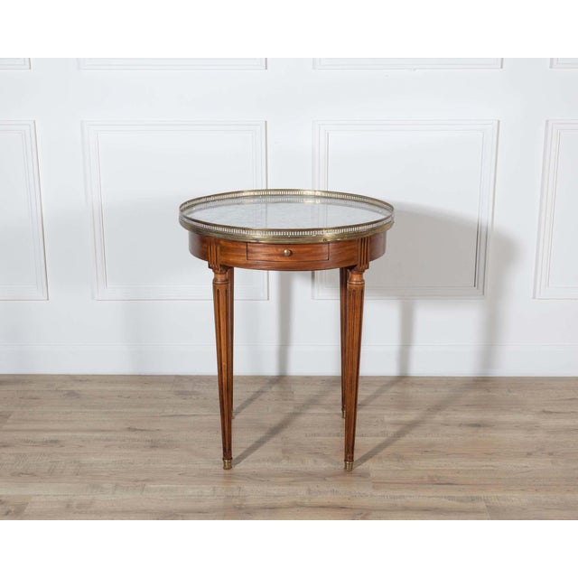 This Louis XVI–style bouillotte table features a circular white marble top bordered by a pierced brass gallery, a defining...