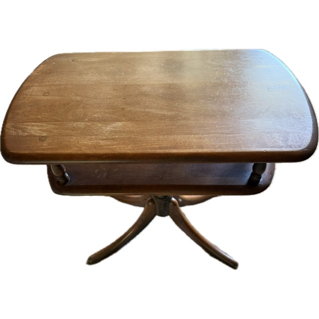 A well-proportioned vintage two-tier pedestal table with a warm reddish wood tone. The floating shelf and slender turned...