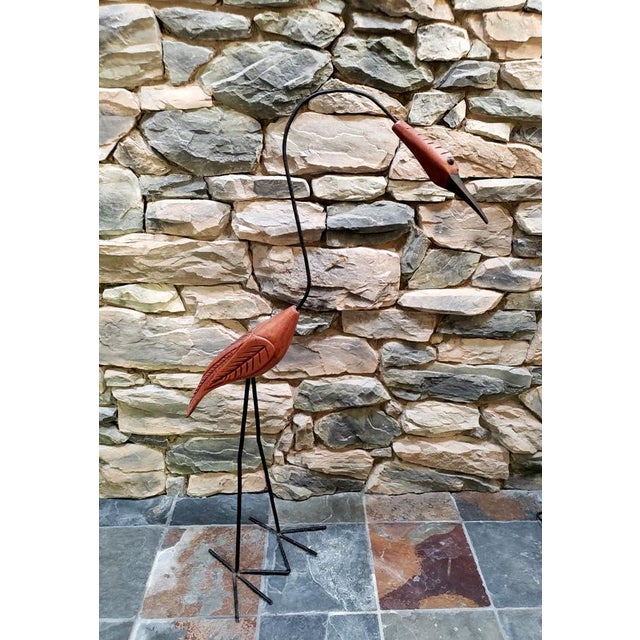 A tall sculptural bird depicting a crane or heron, hand-carved in warm-toned wood and mounted on slender forged iron legs...