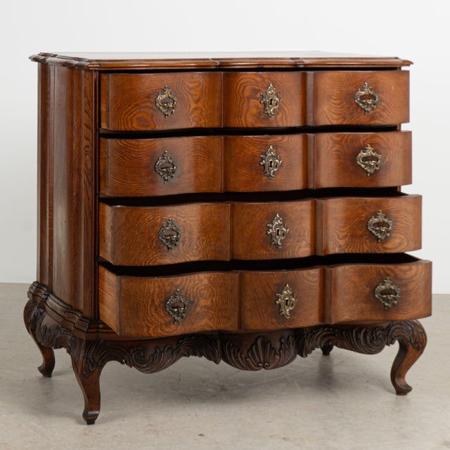 This antique rococo large oak chest of drawers features a serpentine front, brass hardware pulls and is raised on a carved...