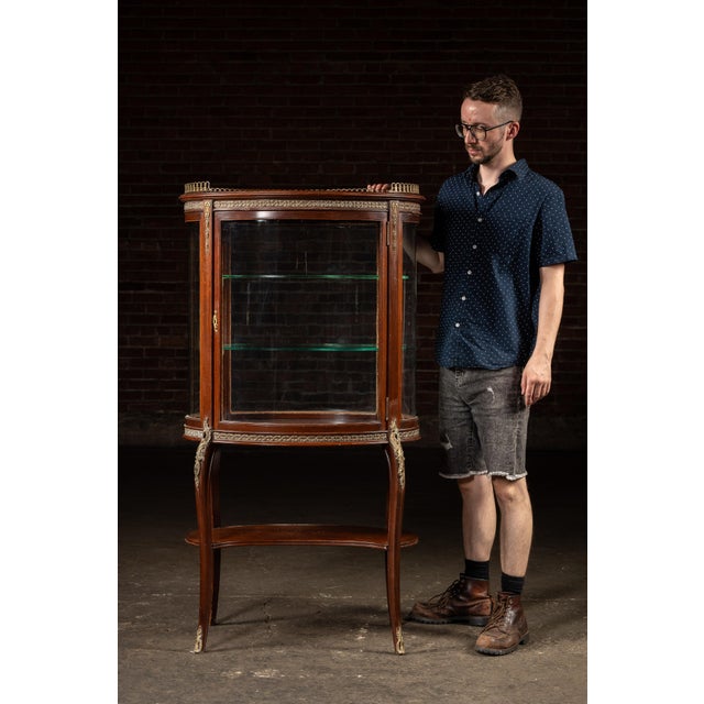 Oval-form mahogany display cabinet with bronze ormolu trim and galleried top. A finely detailed Louis XVI style curio...