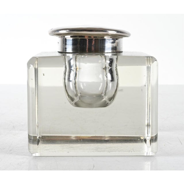 A heavy, square block-form glass inkwell with thick, clear walls and beveled edges. It has a round, domed silver or...