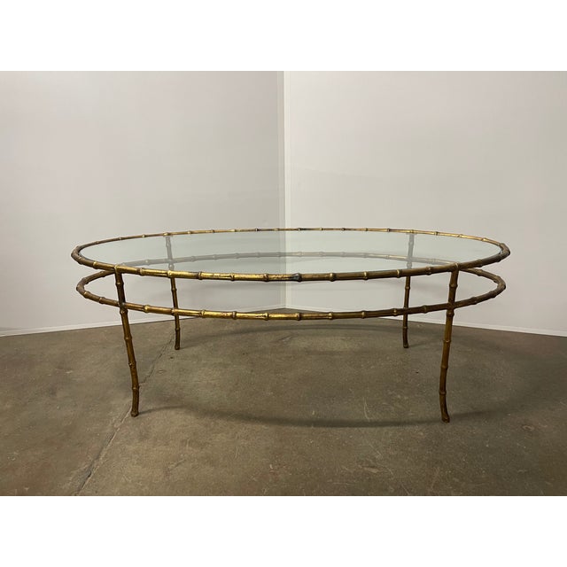 A vintage Hollywood Regency style coffee table with an oval glass top and faux bamboo base in a golden bronze finish....