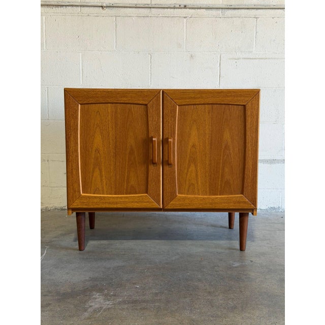 This compact storage unit features a sleek profile accented by framed cabinet doors. It rests gracefully on tapered wooden...