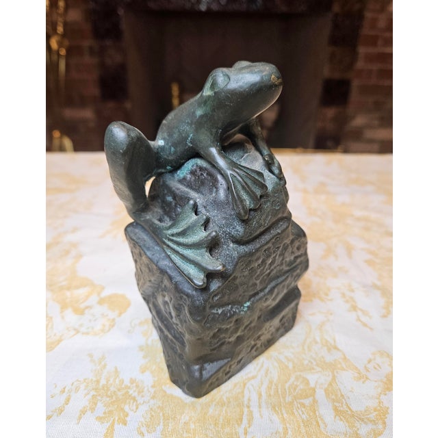 A vintage-style bronze frog sits atop a rock, sculpted in a naturalistic style to resemble either a bullfrog or pond frog...
