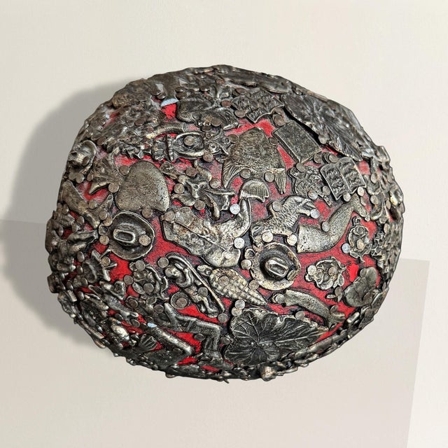 This oblong wooden sphere—lacquered in a sultry, oxblood red—has been entirely armored in silver metal ex-votos, each one...