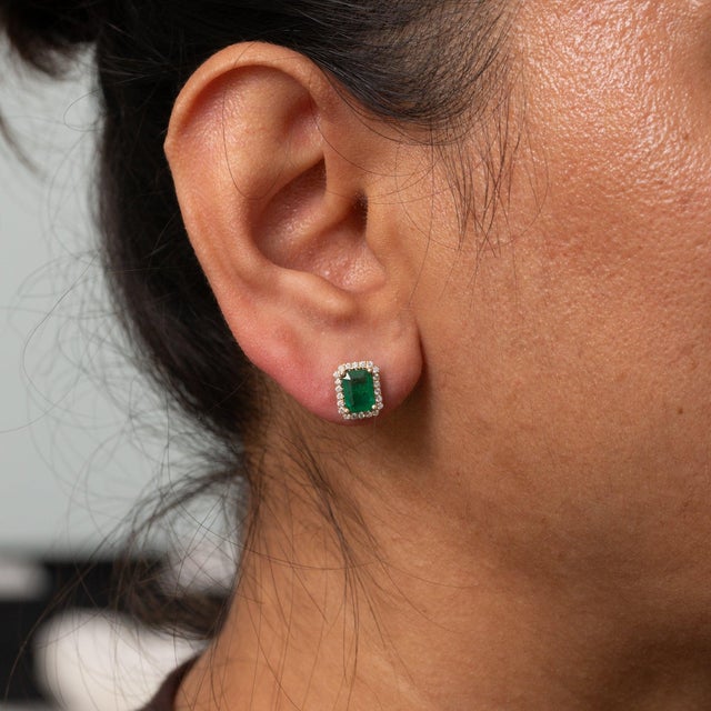 Two emerald-cut zambian emerald stones totalling 2.1 carats glow at the centre of this pair of solid 14K yellow gold...