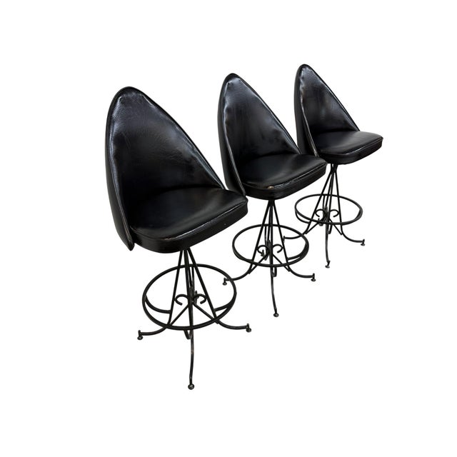 Striking set of three 1970s gothic style swivel bar stools featuring sculptural black vinyl seats and ornate wrought iron...