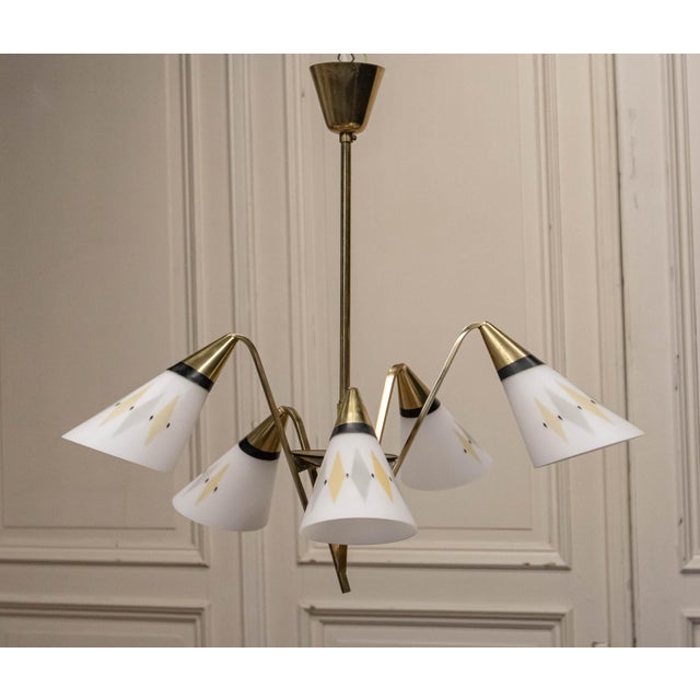 The chandelier features a refined sculptural brass construction with five gracefully angled arms, each supporting a...