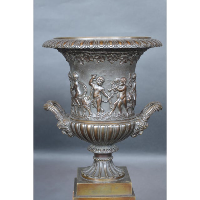 Bronze patinated vases sit on top of a sienna marble base. Cherubs surround the vase, feeding grapes to a woman in a chariot.