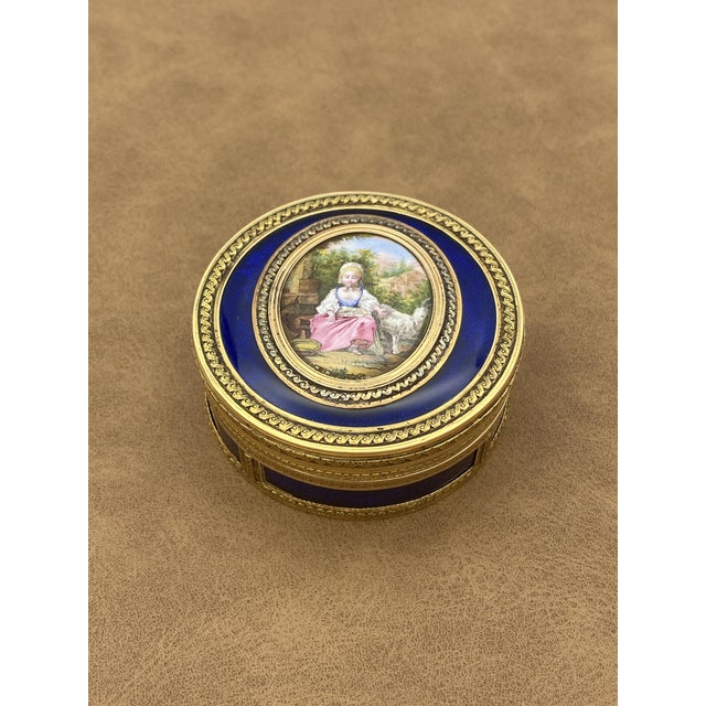 Circular 18K gold snuffbox with a painted scene depicting a young shepherdess seated in a bucolic landscape, with a white...