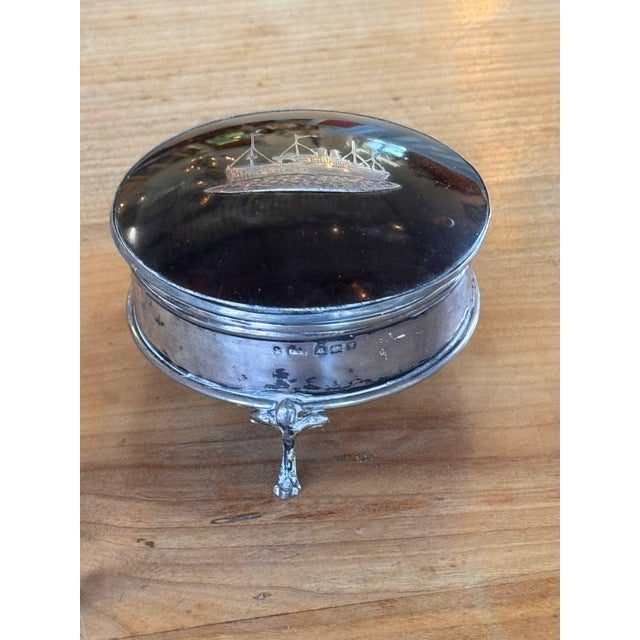 Round box raised on three small shaped feet, the glossy dark lid inlaid with a steamship under full flags, twin funnels...