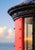 Coastal architectural artwork featuring a vivid red lighthouse exterior contrasted with the warm glow of a Fresnel lens,...