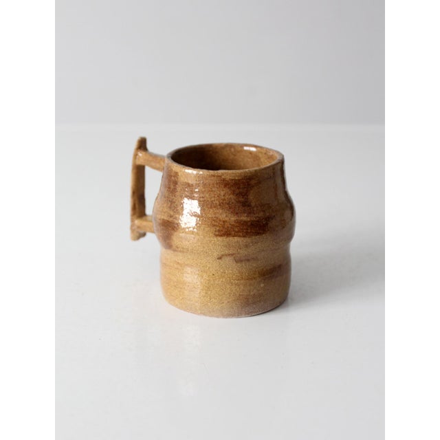 Vintage handmade studio pottery mug featuring warm earth-tone glazes, a sculptural strap handle, and a softly shaped form....