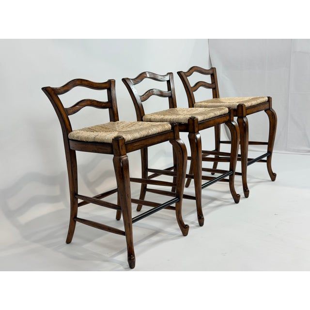 This is a distinguished set of three counter-height stools by Maitland-Smith, crafted in solid wood with a warm, medium-...