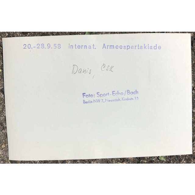 1958 black and white photograph of a gymnast competing in the rings. Printed publication information on the back. Combined...