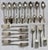 A collection of 26 odd pieces includes: Fifteen Spoons ~ 1.25”W x 6”D x .75”H each Seven Dinner Forks ~ 1”W x 7.25”D x...