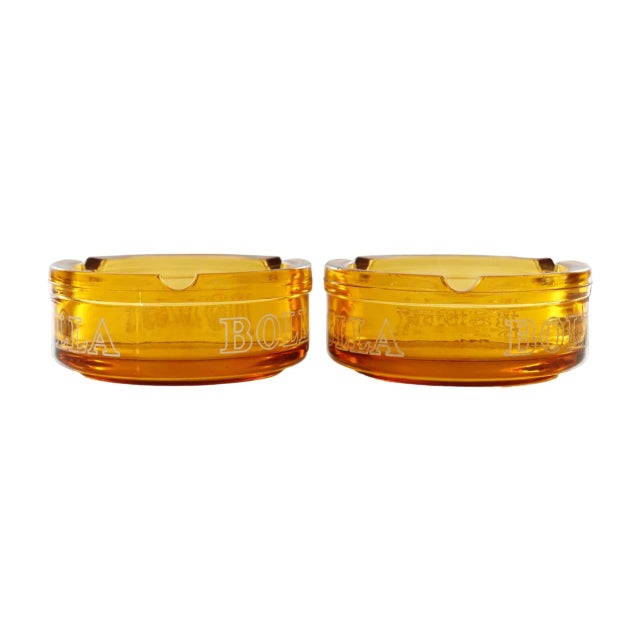A pair of Bolla wine amber glass ashtrays. Each ashtray has three cigarette rests and 'Bolla' in bold white lettering...
