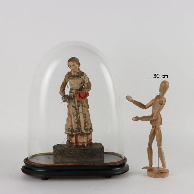 Statue in a display case with terracotta face and limbs and fabric clothes, resting on a wooden base covered in fabric....