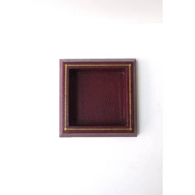 A square red burgundy textured leather jewelry catchall with gold tooling around top edge, by American luxury maker T....