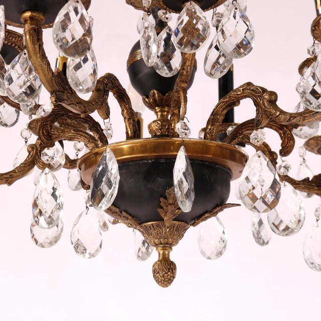 An antique French Empire chandelier offers ebonized and gilt metal frame with scroll and foliate elements, hanging cut...