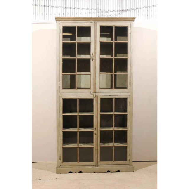 A tall, painted wood glass doors cabinet. This painted wood cabinet from India features a linear profile with four glass...