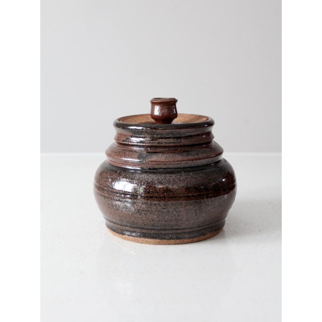 This is a vintage handmade studio pottery jar with a fitted lid. It features a richly variegated brown glaze and a hand-...