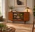 A stylish 1960s mid-century display cabinet in a warm teak finish, featuring central sliding glass doors with adjustable...