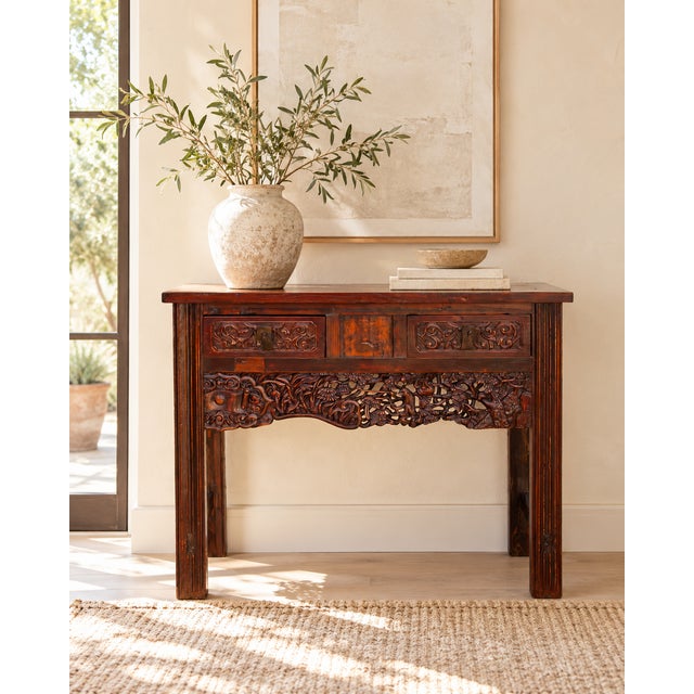 A Chinese altar-style table in warm reddish wood, with a plank top, two drawers and a deeply carved apron. The apron is...